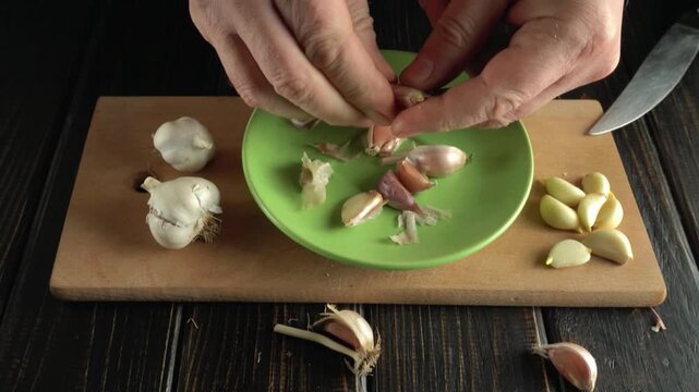 Hands peeling garlic cloves from a green plate on a wooden cutting board with whole garlic bulbs and peeled cloves arranged around