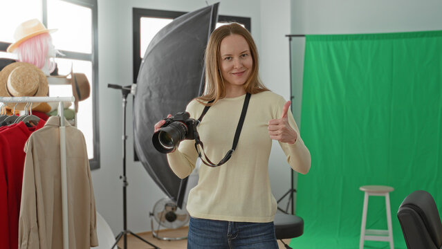Young woman photographer holding dslr camera with strap and giving thumbs up amid green screen, softbox, mannequin head, clothing rack and stool in studio; confidence creativity.