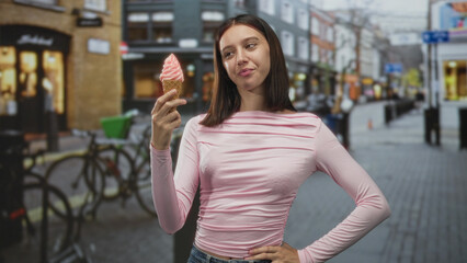 Fototapeta premium Young woman holds pink softserve ice cream cone near face, right hand on hip, smiling on cobblestone shopping street; casual joy.
