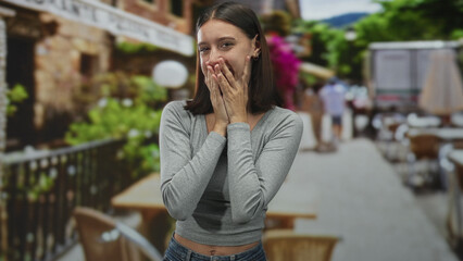 Fototapeta premium Young woman touches cheeks with both hands and smiles in a street cafe by tables and railing; spontaneous joy.