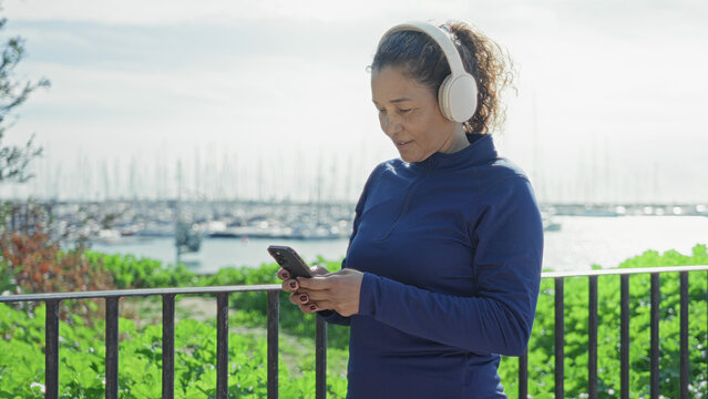 Woman holding smartphone and texting with hands visible on street by a marina railing, wearing headphones and sweatshirt; calm mindfulness.
