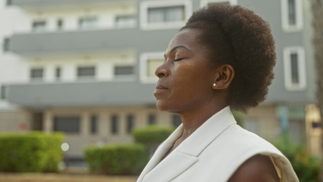 Woman with closed eyes tilting head upward, showing earring and neck, standing near modern building facade; calm reflection.