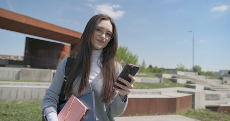 A hand-held shot of a young female student in casual clothes wearing glasses holding a notepad and laptop in her hands and using a mobile phone while standing in an urban park on a summer day © Anatoly Repin