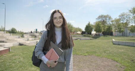 A hand-held shot of a young female student in casual clothes, wearing glasses and a backpack, holding a notebook and laptop in her hands, walking through an urban park on a summer day. © Anatoly Repin