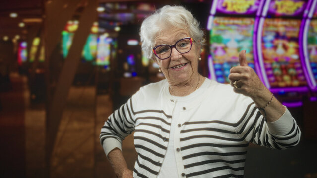 Elderly woman raises fist at a casino slot machine, smiling with glasses, striped cardigan, ring and bracelet visible; victory luck celebration.
