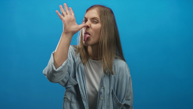 Young blonde woman wearing denim shirt with tongue out making thumb to nose gesture in a bright blue studio; playful teasing.