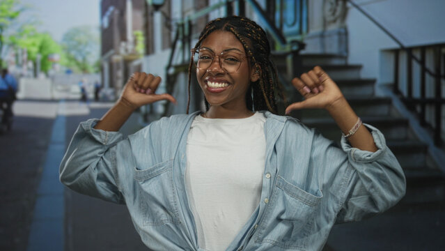 Woman with glasses and braided hair, thumbs pointing to chest on a city street by stone staircase; confidence pride.