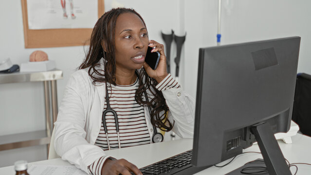 Woman doctor holding smartphone to ear and typing at computer desk with stethoscope visible in clinic building; care coordination concern.