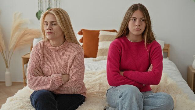Woman and daughter sitting on bed with crossed arms, seated apart and avoiding eye contact in bedroom domestic setting; family tension.