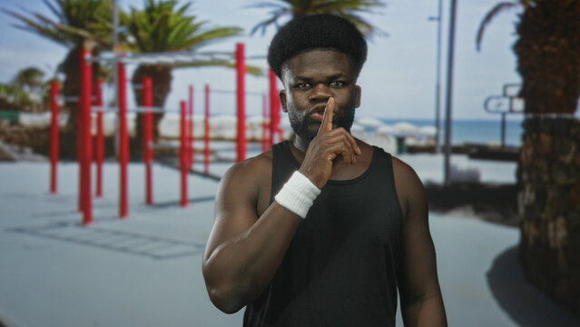 Man with finger to lips for silence at outdoor street gym by red pullup bars and palm trees, wearing black tank and wristband; quiet determination.