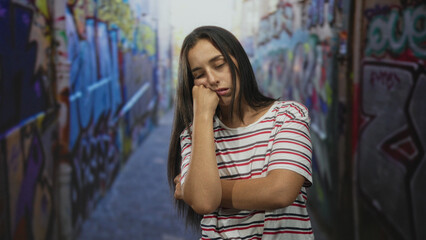 Obraz premium Woman resting chin on fist in graffiti street alley, wearing striped tshirt with closed eyes and long hair, freckle visible; tired solitude.