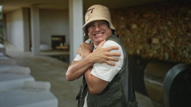 Man hugging shoulders and grimacing in a building pool area while wearing a fishing vest and bucket hat; discomfort solitude.