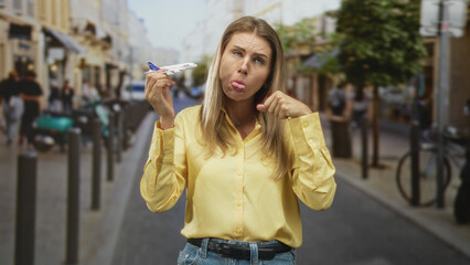 Fototapeta premium Woman holding a toy airplane in her right hand, left fist by chin, pouting expression on a busy street; travel frustration.