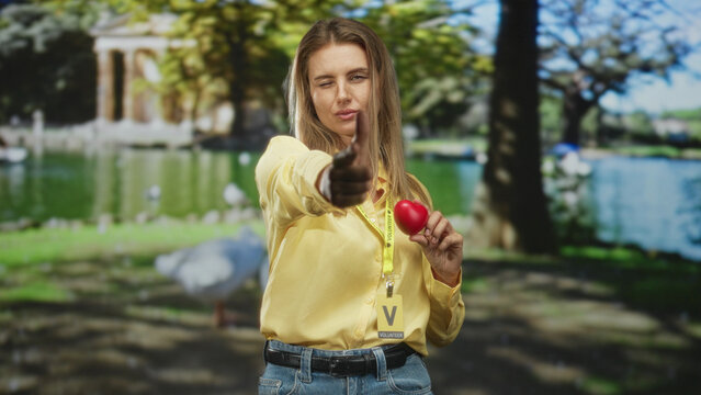 Woman points finger at camera while holding red heart and visitor badge in park; compassion volunteer care.