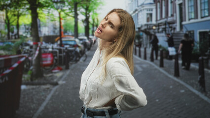 Fototapeta premium Woman in white blouse with hand on lower back for backache on city street amid bicycles and bollards; discomfort everyday strain.