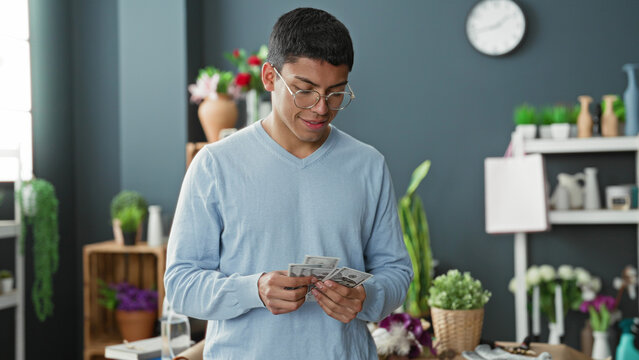 Man florist counts cash in hands amid vases, flowers and potted plants in studio, smiling while tallying dollars; quiet satisfaction.