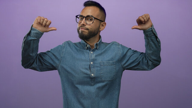 Young man with glasses and thumbs pointing to chest in studio with purple backdrop; confidence self belief.
