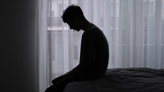 Silhouette man seated at curtained window, tired office worker with head bowed and hands on knees, dim evening light and gray backlight, small urban studio