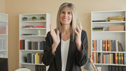 Woman clapping hands and smiling, middleaged blonde in black blazer before bookshelves in office building  warm encouragement. © Krakenimages.com