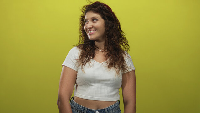 Woman smiling with bare midriff wearing white tshirt and jeans three quarter view in lime green studio; confidence casual natural.