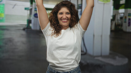 Woman in white t shirt raising arms and smiling with clenched fists at petrol station forecourt by fuel pumps and columns  joy victory celebration. © Krakenimages.com
