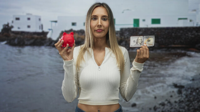 Woman holds red piggybank and hundred dollar banknote at seaside with closed eyes and visible navel piercing; indecision saving spending.