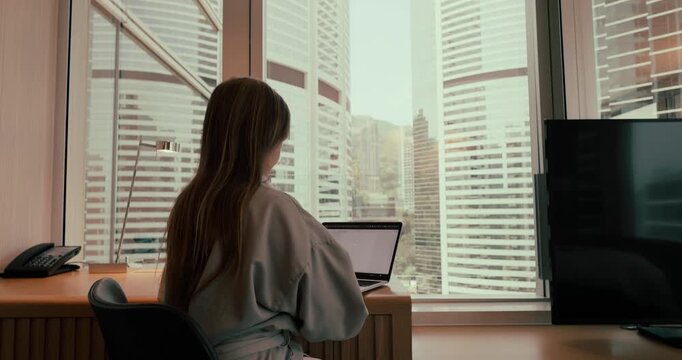 Woman working on laptop at desk in high-rise office or hotel room overlooking skyscrapers in Hong Kong during daytime. Slow zoom in from behind. Slow motion.