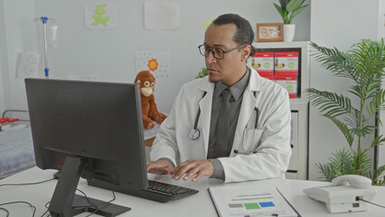 Naklejka premium Man doctor types on keyboard while reviewing charts at a computer in a pediatric exam room building; focused care documentation.