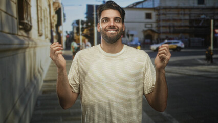 Naklejka premium Hispanic man with beard clapping hands and smiling on a sunlit street beside a building, wearing a striped t shirt while people and a taxi appear behind him; joy gratitude.