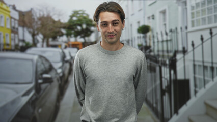 Obraz premium Man in grey sweater points finger with visible hand on a residential street beside parked cars and iron railings; casual confidence.