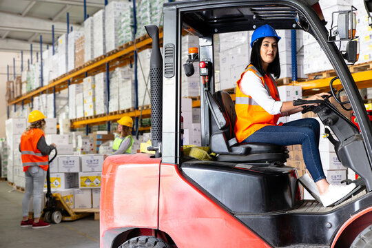 In a warehouse, a woman wearing a blue hard hat and vest operates a forklift while two colleagues in yellow hard hats and vests stack boxes on pallets, showcasing warehouse operations and teamwork.