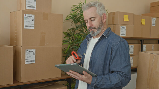 Man scans parcel with barcode scanner while holding tablet amid stacked boxes on shelf in warehouse; focused efficiency.