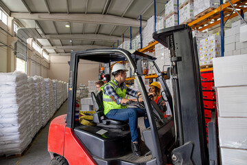 A warehouse worker navigates a forklift through a warehouse, surrounded by stacked pallets and shelves. He wears a yellow safety vest and a hard hat. © bulentbaris