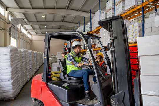 A warehouse worker navigates a forklift through a warehouse, surrounded by stacked pallets and shelves. He wears a yellow safety vest and a hard hat.