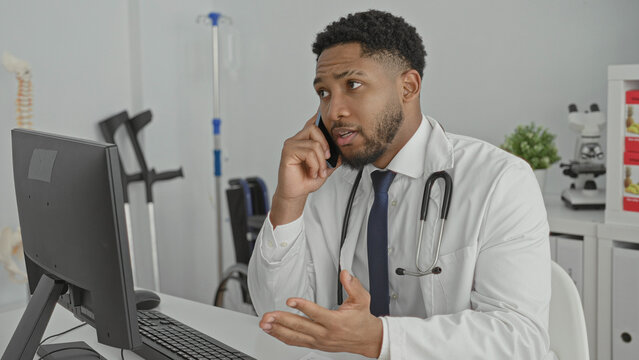 Young man doctor with stethoscope holding phone to ear and gesturing toward computer in clinic; concerned professionalism.