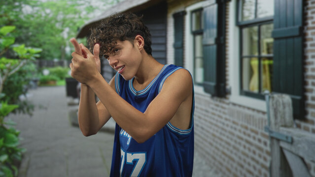 Teenage boy in blue los angeles basketball jersey clapping hands and miming finger gun on a street; playful swagger.