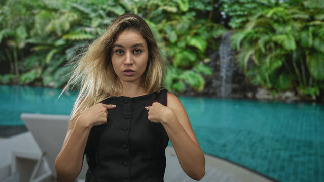 Woman points finger to chest with palms up beside turquoise pool in resort building, black dress and windblown blonde hair; confusion self doubt.