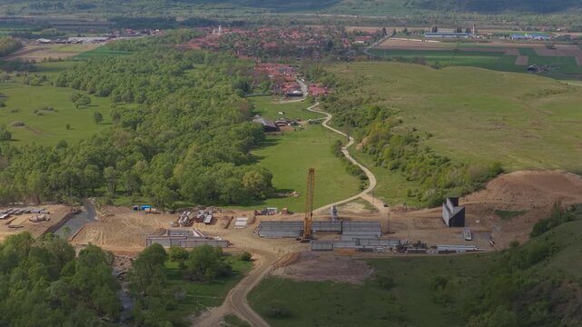 Dynamic highway construction site with a yellow crane building a bridge over a river in the picturesque rural landscape of Sibiu Fagaras, high altitude aerial lateral tracking shot 4K