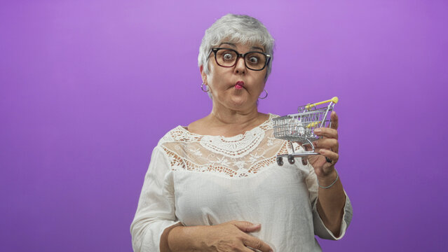 Senior woman holding a small shopping cart with both hands, puckered lips and raised eyebrows in a purple studio, inspecting the toy cart; playful surprise.