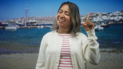 Fototapeta premium Hispanic middleaged woman pointing index finger up, smiling and wearing white cardigan and striped shirt at a harbor; joy playfulness.