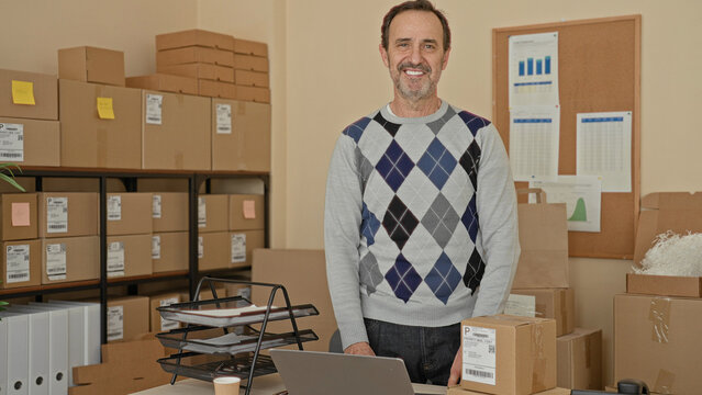 Man smiling while packing cardboard boxes and leaning over a laptop with hands on desk in a building parcel office; small business pride.