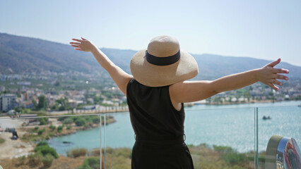 Fototapeta premium Woman with outstretched arms and straw sunhat on building deck overlooking turquoise sea and coastal town, facing away from camera; serenity escape.