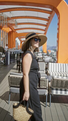 Fototapeta premium Woman smiling, hand on glass railing on building terrace of cruise deck with sunhat and sunglasses, wearing black dress; serenity.