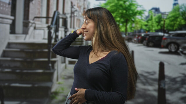 Young woman pointing with hand and miming phone gesture on street, wearing necklace and dark vneck, long hair, smiling sideways; playful smile joy.