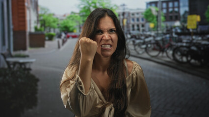 Fototapeta premium Woman with clenched fist raised at camera on city street with bicycles and brick buildings visible; anger defiance resilience.