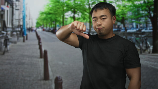 Man in black t shirt showing thumbs down with hand on street lined with bicycles and green trees; disapproval.