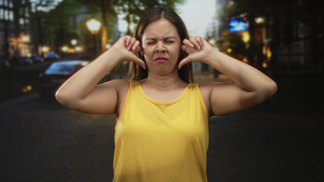 Woman plugs ears with fingers and eyes closed on busy city street at dusk wearing yellow tank top and necklace; annoyance urban noise.