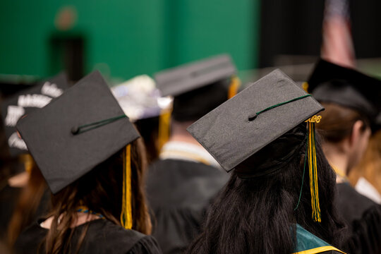 A group of graduates in black gowns and mortarboards with gold tassels are seen from behind at an indoor commencement ceremony, a green gymnasium backdrop behind them.