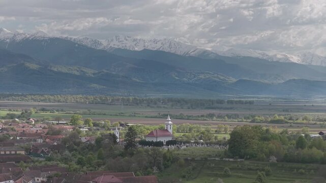 Peaceful Ucea de Sus village with a white church at the foot of the snow capped Fagaras mountains, scenic aerial view with slow descent ungraded log 4K