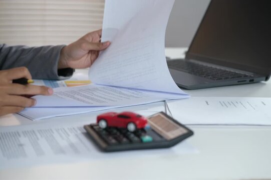 An insurance underwriting officer who handles documents, checks documents. Working at a desk with a laptop, pointing to a document with a picture of a small red car on it.
